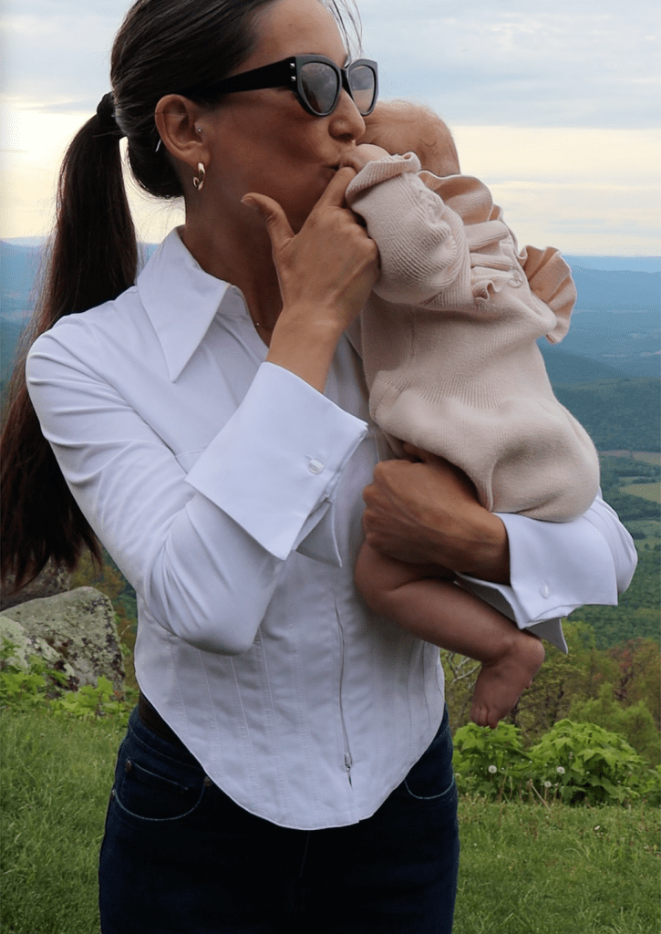 A mother wearing a white blouse kisses her baby while standing outdoors, with mountains in the background.
