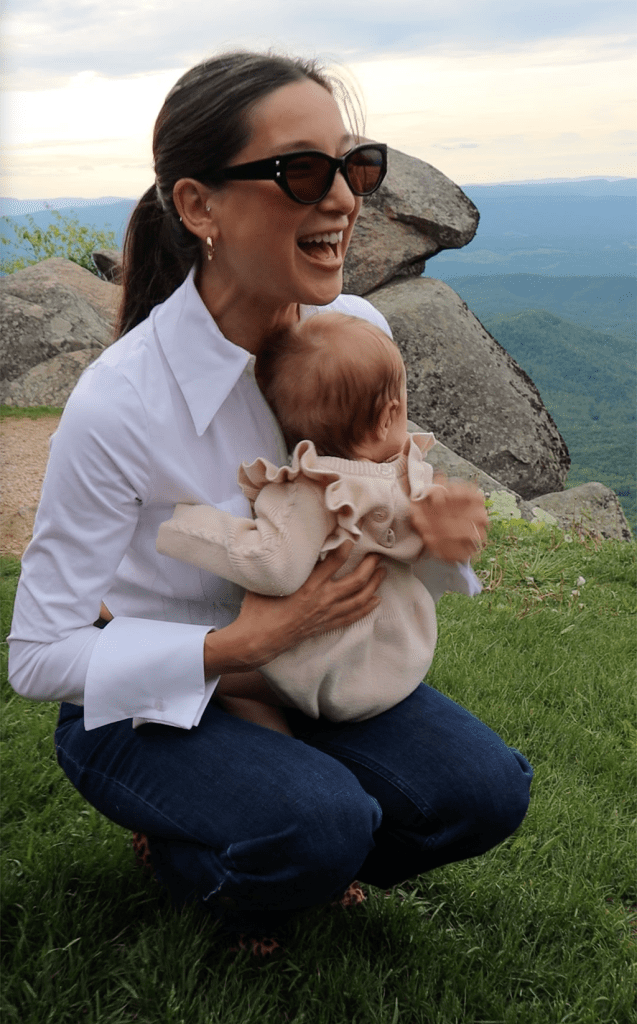 A smiling woman wearing sunglasses and a white shirt is holding a baby in a cozy outfit, crouching on grassy land with a mountain view in the background.