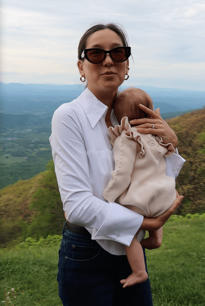 A woman wearing sunglasses holds a baby while standing on a grassy hillside, with mountains visible in the background.
