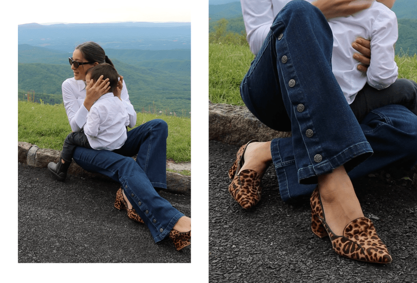 A mother lovingly embraces her child while sitting outdoors, surrounded by a scenic mountain backdrop. She wears a white blouse and stylish dark blue jeans featuring button details, along with leopard print heels. The moment captures the essence of warmth and connection in motherhood.