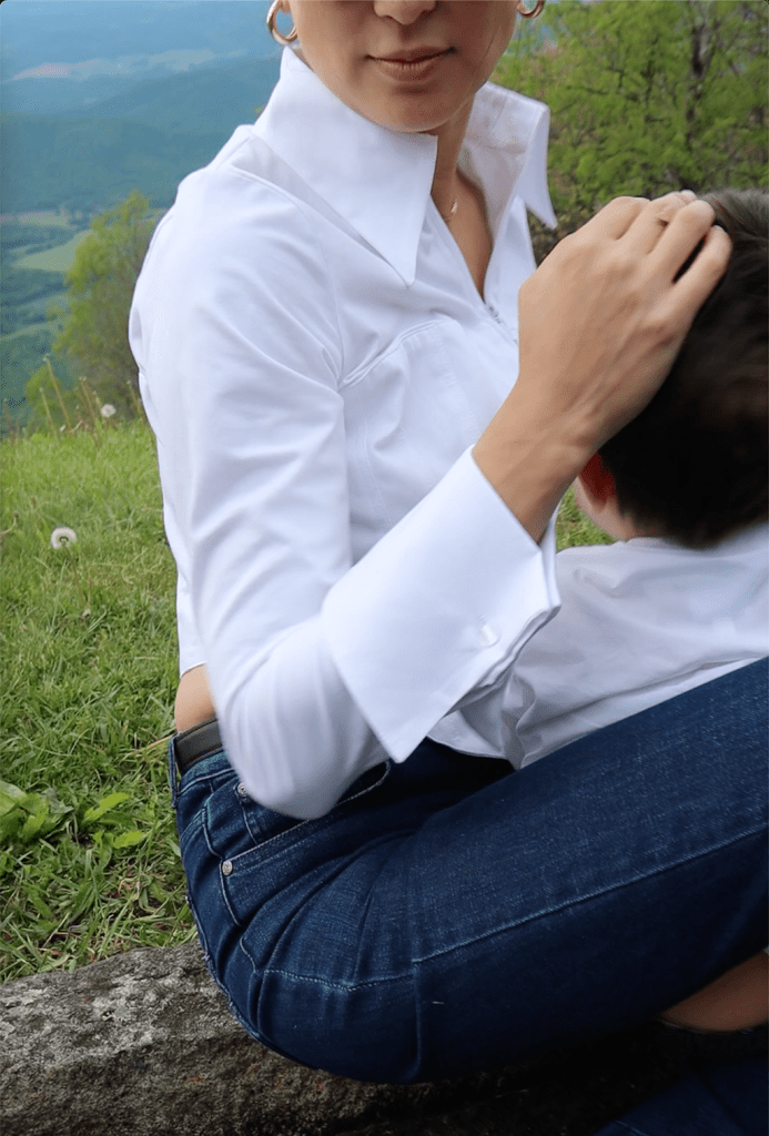 A woman wearing a crisp white shirt and jeans, sitting outdoors with a child in her lap, surrounded by green grass and a scenic mountain view.