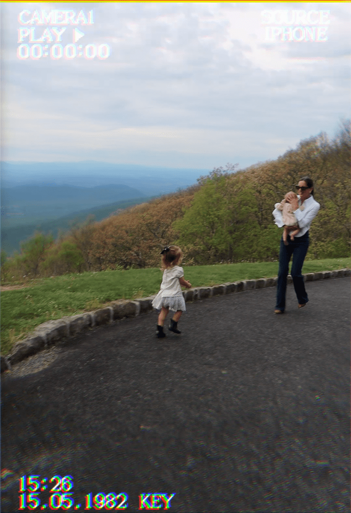 A mother holds a baby while walking on a scenic path, as a young child in a white dress runs playfully nearby, surrounded by a lush green landscape and cloudy sky.