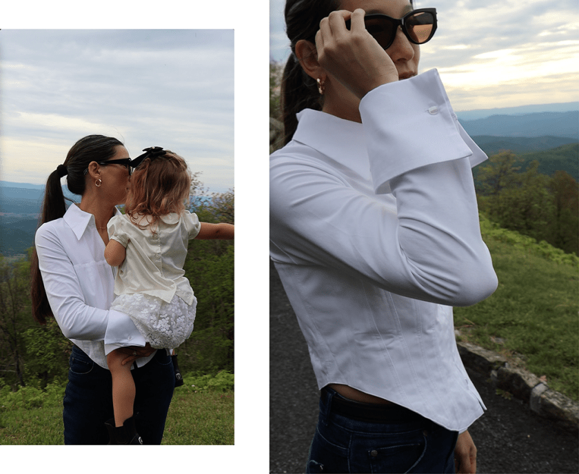 A woman holding a child while wearing a white shirt, standing against a scenic mountainous backdrop.