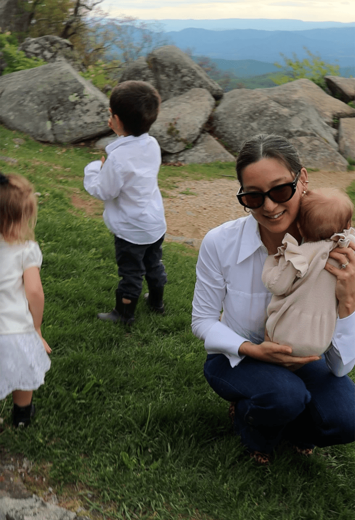 A mother holding her baby while smiling, with two young children playing nearby on a grassy field with rocks and mountains in the background.