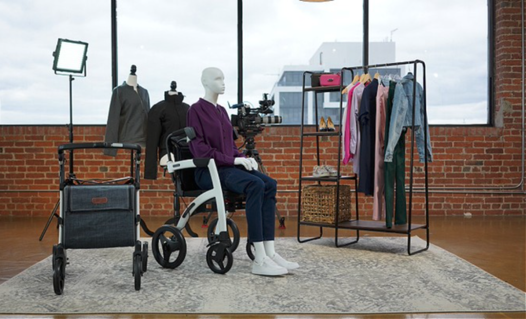 Molly Farrell, a white woman with brown hair, is shown in this photo wearing ULEX, one of the brands Tracy designed and helped launch. Molly is wearing a royal blue wrap cardigan and gray pants, while seated on bleachers. She is smiling brightly and her pink forearm crutches are visible in the photo. 