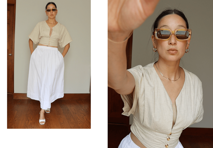 A person wearing a beige crop top with buttons and a white, flowy skirt, posing in front of a neutral backdrop while holding a camera with oversized sunglasses on.
