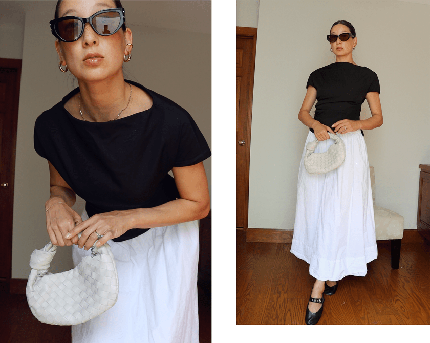A person wearing a fitted black top and a flowing white skirt, holding a small woven handbag in a stylish pose. The background features a wooden door and a chair.