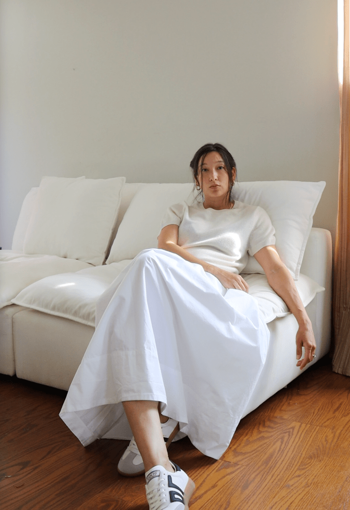 A woman sitting on a sofa, wearing a Quince light sweater and a long white skirt, with sneakers on her feet, in a well-lit indoor space.
