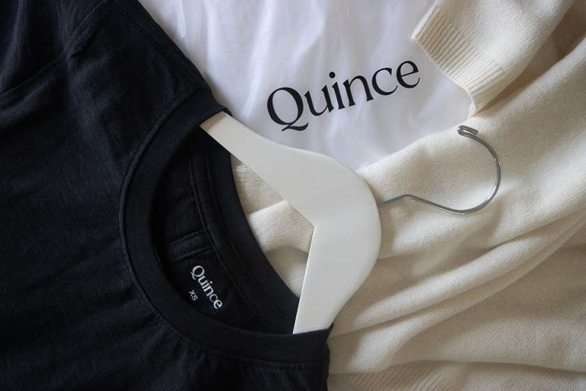 Close-up of a black Quince t-shirt and a white hangtag showcasing the Quince logo, with a cream cashmere sweater in the background.