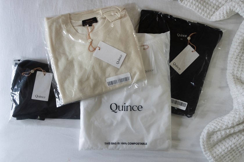 A selection of Quince clothing items including a beige sweater and two black garments, all in clear packaging with tags on a white background.