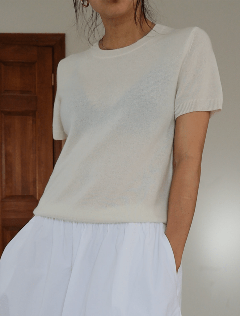 Model wearing a light cashmere sweater and a white skirt, standing indoors near a wooden door.