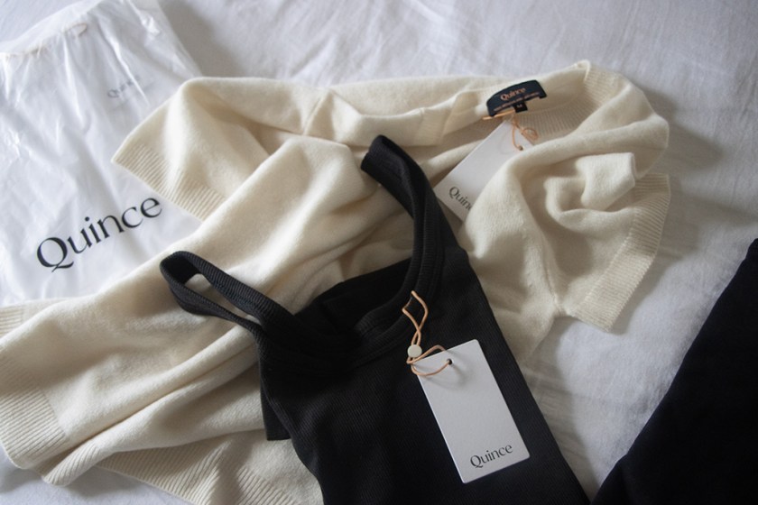 A flat lay of clothing items from Quince, featuring a cream sweater, a black tank top, and a white shopping bag with the Quince logo.