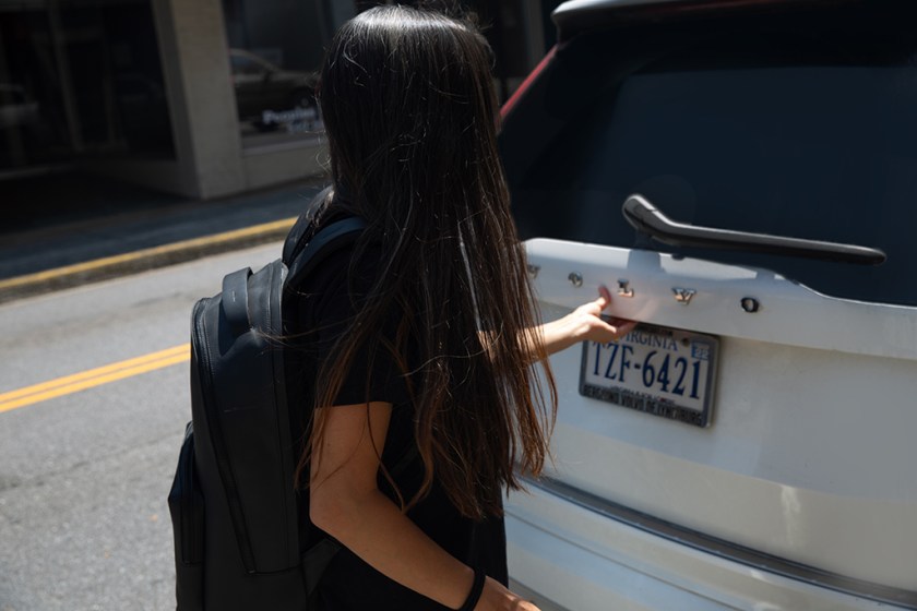 A person with long hair is standing beside a parked vehicle, wearing a sporty black backpack and reaching towards the car's hatchback.