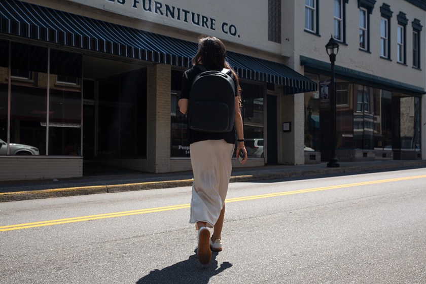 A woman walking on a city street wearing a sporty backpack, dressed in a fitted black top and light-colored skirt, with buildings and a storefront in the background.