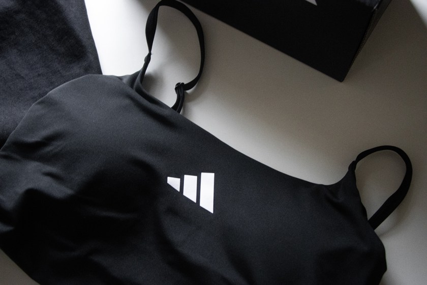 Black sports bra with thin straps, featuring a white adidas logo, placed on a light surface beside dark fabric.