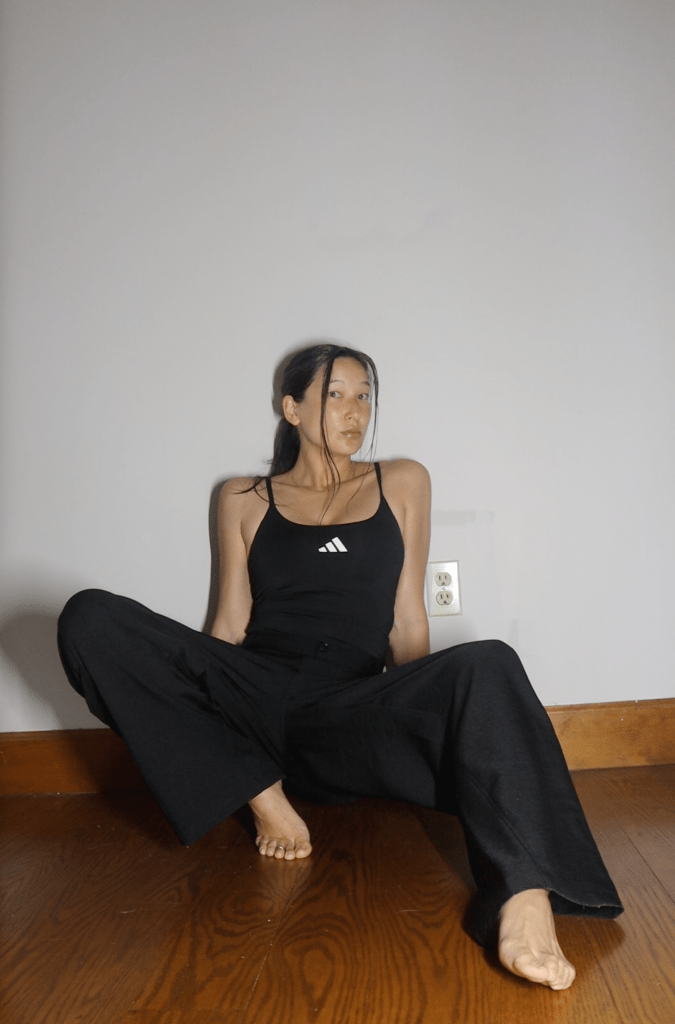 A person sitting on the floor in a black tank top and wide-leg black pants, with a minimalist background.