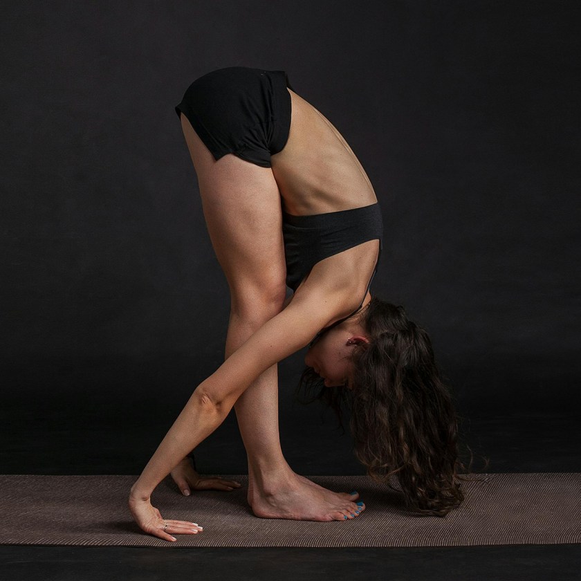 Person performing a forward bend yoga pose on a mat, wearing a black sports bra and shorts, against a dark background.